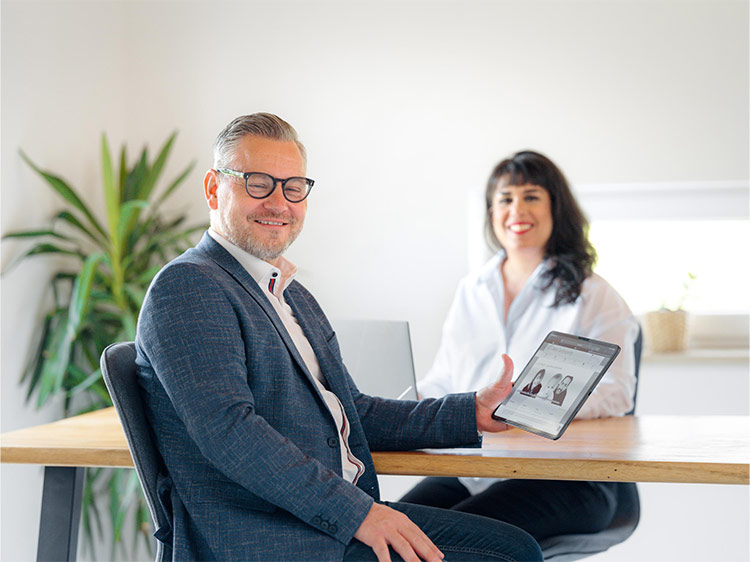 Lukas Senicky zeigt am Tablet Inhalte von GT Capital, während Monique Senicky im Hintergrund lächelt – authentische Versicherungsberatung in moderner Arbeitsatmosphäre.