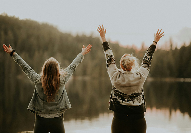 Zwei Frauen stehen mit dem Rücken zur Kamera vor einem See im Wald und strecken fröhlich die Arme in die Luft dank Absicherung durch GT Capital