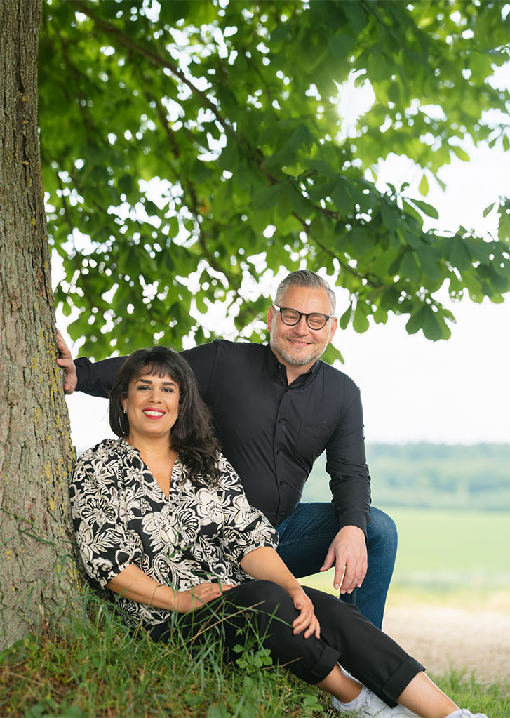 Monique und Lukas sitzen neben einem Baum und schauen freundlich in die Kamera.